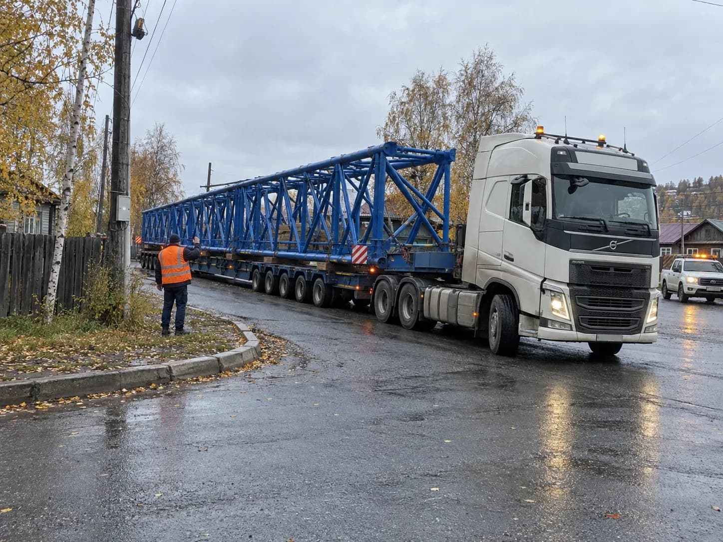 Тягач с длинной секцией вышки проходит поворот, на углу рабочий подаёт знаки водителю